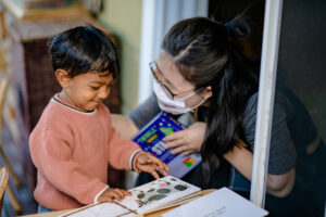 A young child reads a book with an adult wearing glasses and a face mask beside them, smiling.
