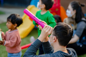 Children wearing masks play outside; one child looks through a pink tube near a slide, others watch nearby.
