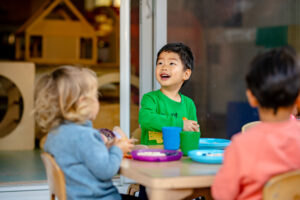 Three young children sit at a table in a classroom, eating and talking together.