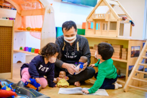 An adult and two children wearing masks play with dolls and toys in a classroom or playroom setting.