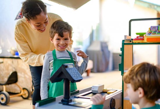 A smiling boy in a green apron stands at a toy cash register, watched by an adult and another child.