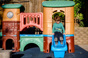 A young boy slides down a blue slide on a colorful plastic playground set in a sunny outdoor area.