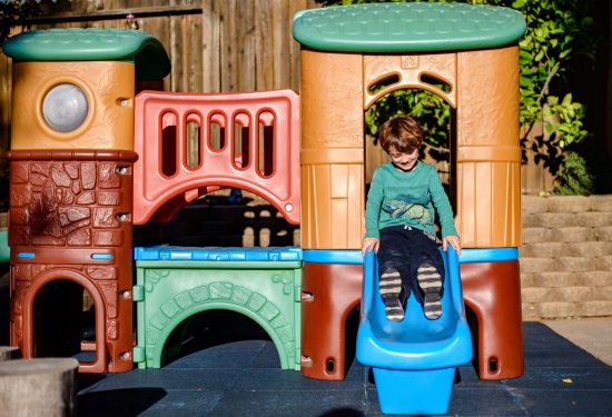 A young boy slides down a blue slide on a colorful plastic playground set in a sunny outdoor area.