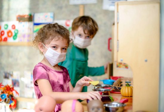 Two young children wearing masks play with toy kitchen items in a colorful classroom.