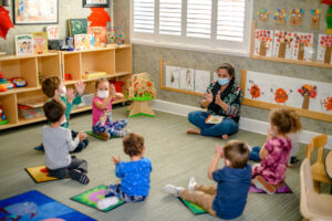 Teacher and young children sit in a circle on mats in a classroom, clapping and wearing face masks.