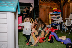 A woman kneels beside a young boy opening a playhouse door in a backyard with toys and outdoor furniture.