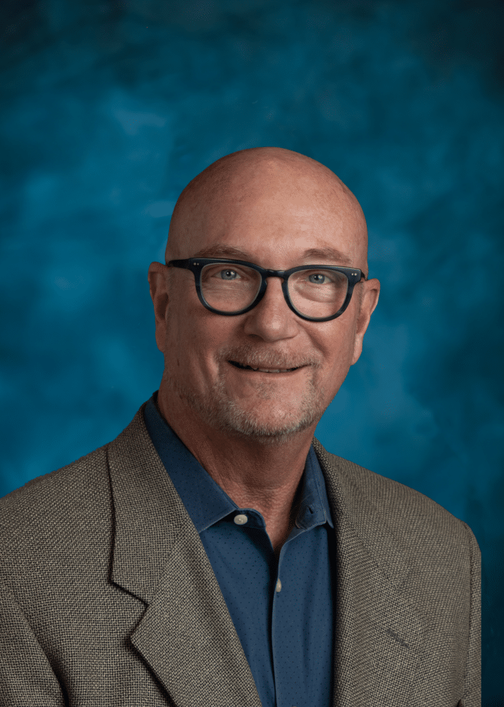 Bald man with glasses and a goatee, wearing a tan blazer over a blue shirt, poses in front of a blue backdrop.