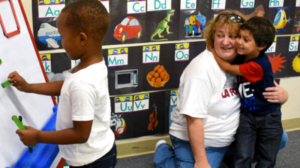 A young boy hugs a teacher while another child plays with magnetic letters in a classroom.