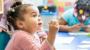 Young girl with braids and beads holding a pencil, sitting at a table, engaged in classroom activity.