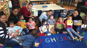 Adults and children sit on a colorful alphabet rug, holding picture books and smiling in a classroom setting.