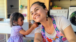 Smiling woman in a colorful apron with a young girl indoors near a washing machine and wooden shelves.