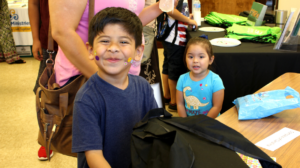 Smiling boy with stickers on his face stands by a table, with a girl in blue shirt behind him indoors.