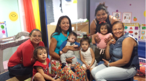 Five women and four young children smiling together in a colorful playroom with educational posters on the wall.