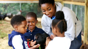 A woman and three children smile and gently pet a black chicken at an outdoor enclosure.