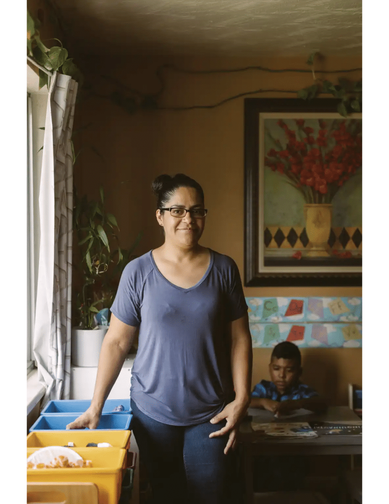 A woman stands by bins in a cozy room while a child sits at a table in the background.