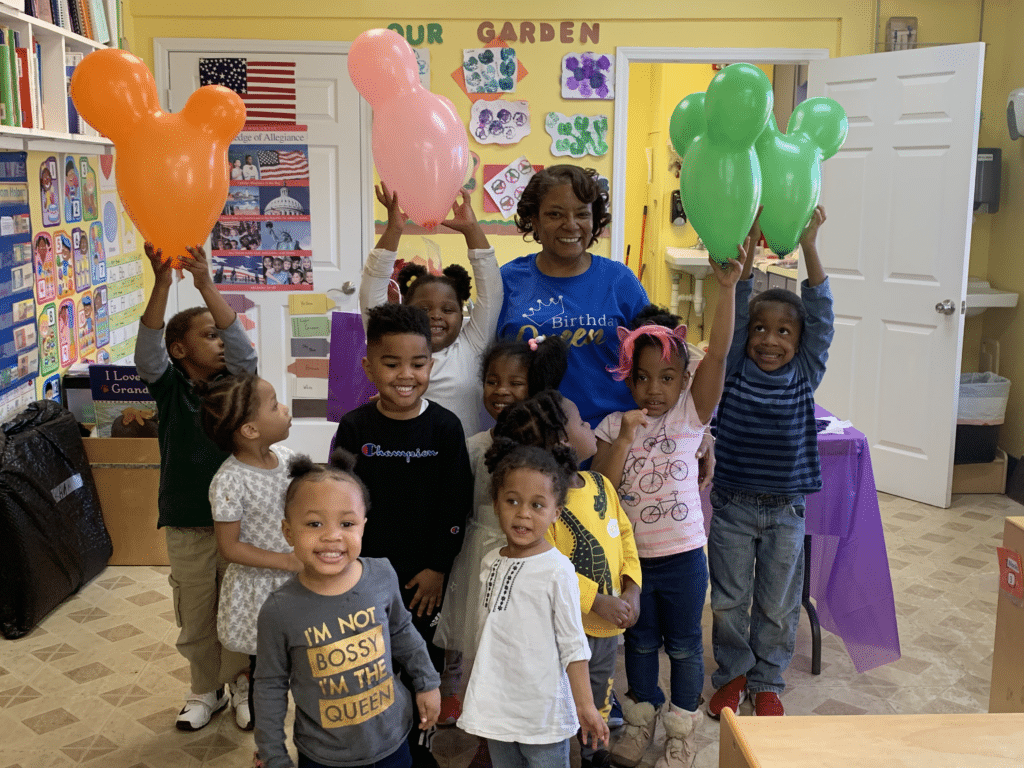 A group of smiling children and an adult hold colorful balloon animals in a classroom decorated for a party.