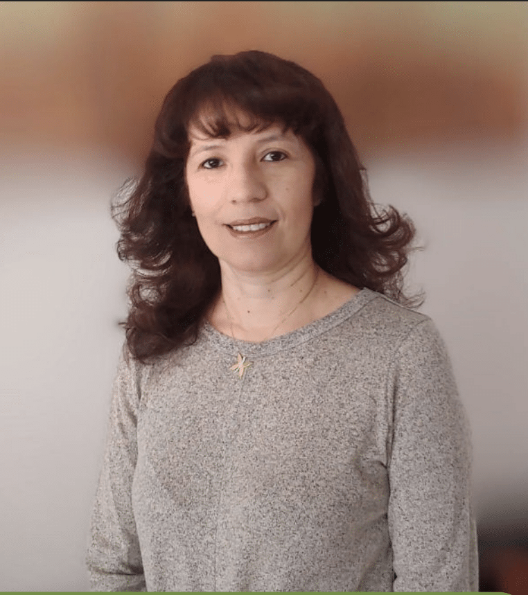 Woman with wavy brown hair wearing a grey sweater, smiling, standing in front of a blurred background.