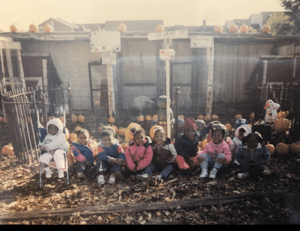 A group of children in jackets sits on autumn leaves at a pumpkin patch in front of a shed.