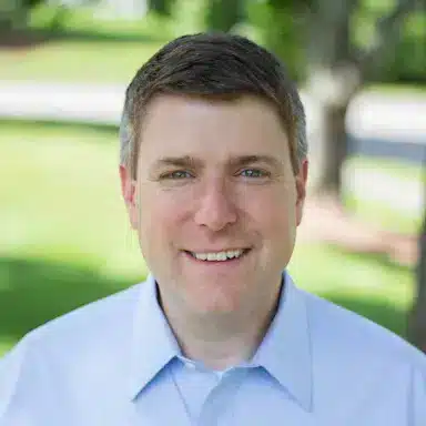 A man with short brown hair smiles outdoors, wearing a light blue collared shirt; trees in the background.